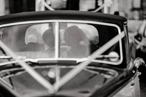Black and white photo of a couple sharing a kiss inside a vintage classic car, captured through the windshield.