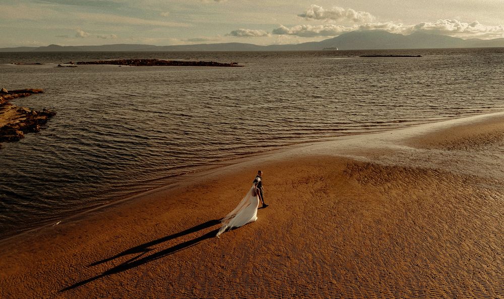 Aerial view of a bride in a flowing white dress walking along a sandy beach shore at sunset.
