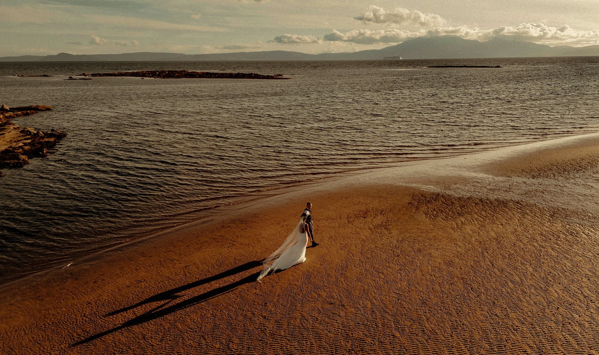 Aerial view of a bride in a flowing white dress walking along a sandy beach shore at sunset.