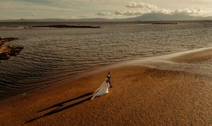 Aerial view of a bride in a flowing white dress walking along a sandy beach shore at sunset.