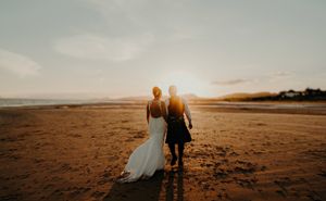 Couple in wedding attire walking hand-in-hand on a sandy beach at golden sunset.