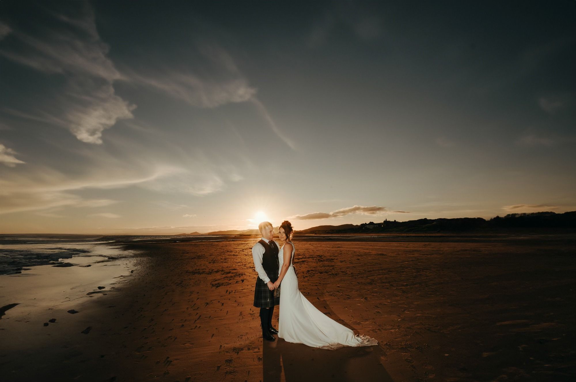 Bride and groom share a romantic sunset kiss on a sandy beach, with golden light reflecting on the shore.