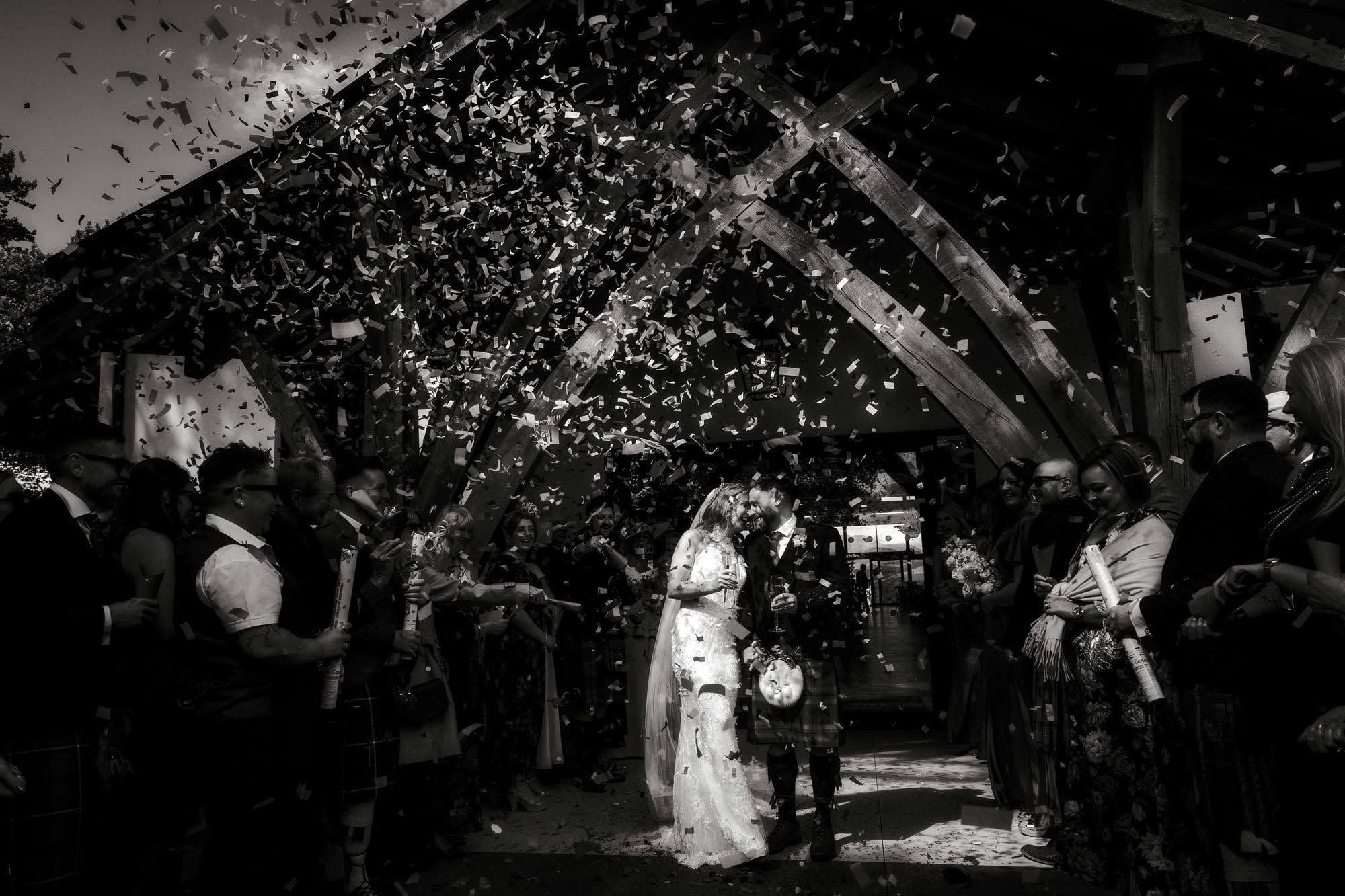 Black and white wedding photo of bride and groom walking under confetti shower surrounded by cheering guests.