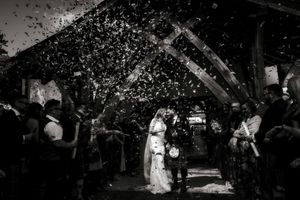 Black and white wedding photo of bride and groom walking under confetti shower surrounded by cheering guests.