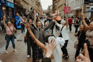 Joyful wedding celebration on a busy city street with guests cheering and raising hands around a bride in a white dress.