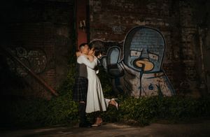Newlywed couple sharing a romantic kiss against a brick wall with colorful graffiti art at their wedding.