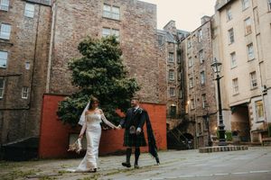 Bride and groom walk hand in hand through a historic Edinburgh courtyard, groom wearing traditional Scottish kilt.