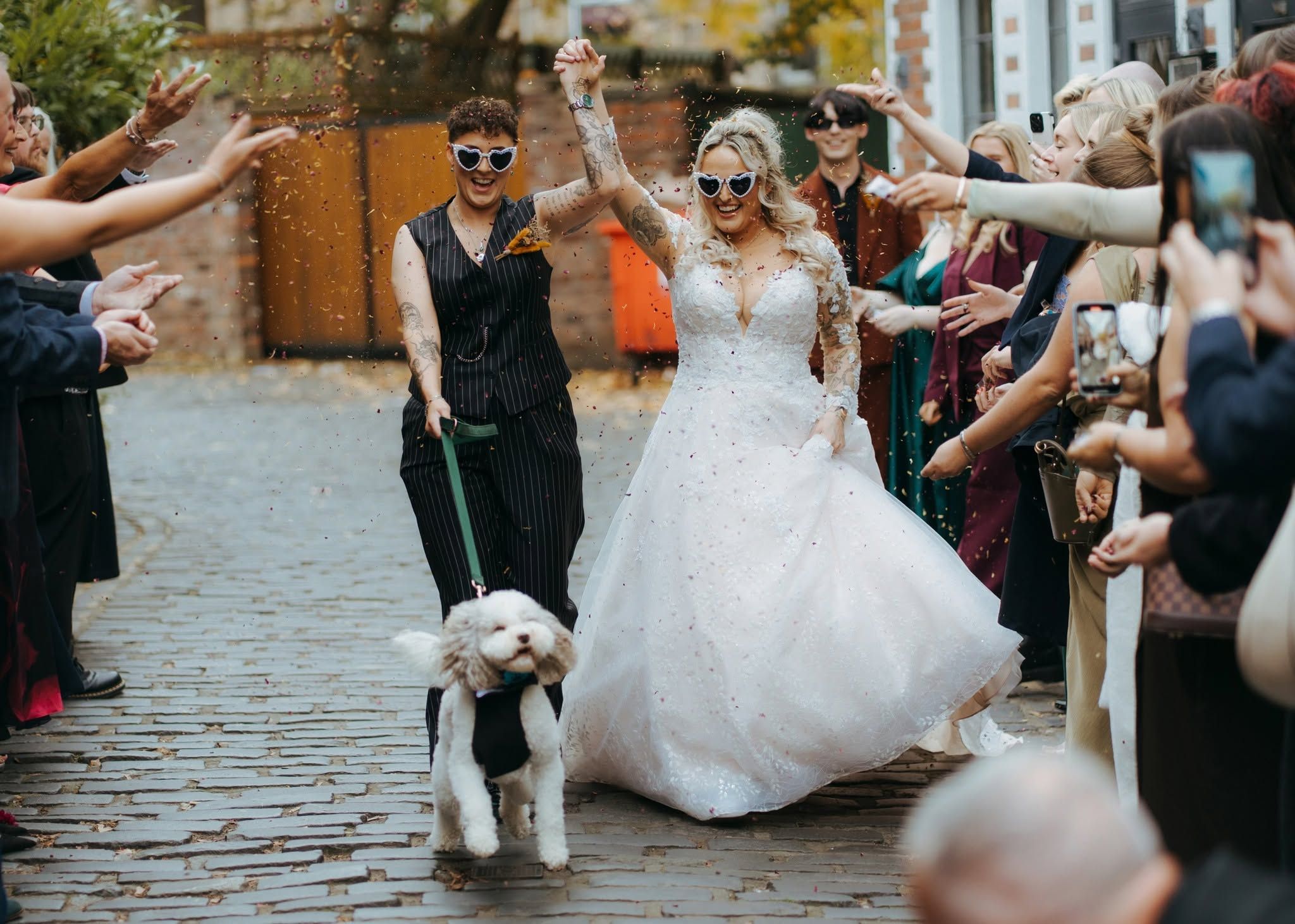 Bride in white lace gown walks with fluffy dog in tuxedo through cheering guests at outdoor wedding celebration.