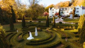 Aerial view of a bride and groom posing in a manicured garden maze beside an elegant white manor house in autumn.