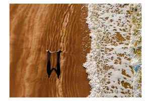 Aerial view of two people holding hands on a sandy beach with ocean waves washing ashore.