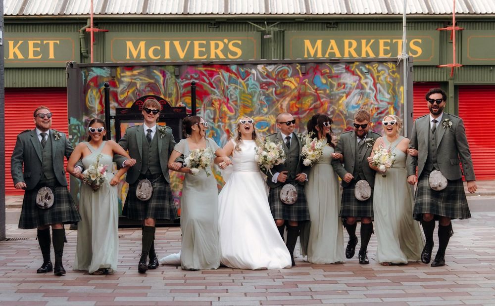 Wedding party in kilts and gray gowns posing outside McIver's Markets, with bride and groom centered.
