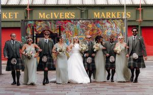 Wedding party in kilts and gray gowns posing outside McIver's Markets, with bride and groom centered.