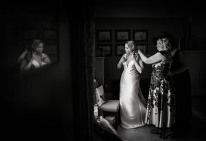 Black and white photo of a bride being helped into her wedding dress by two women in an elegant room.