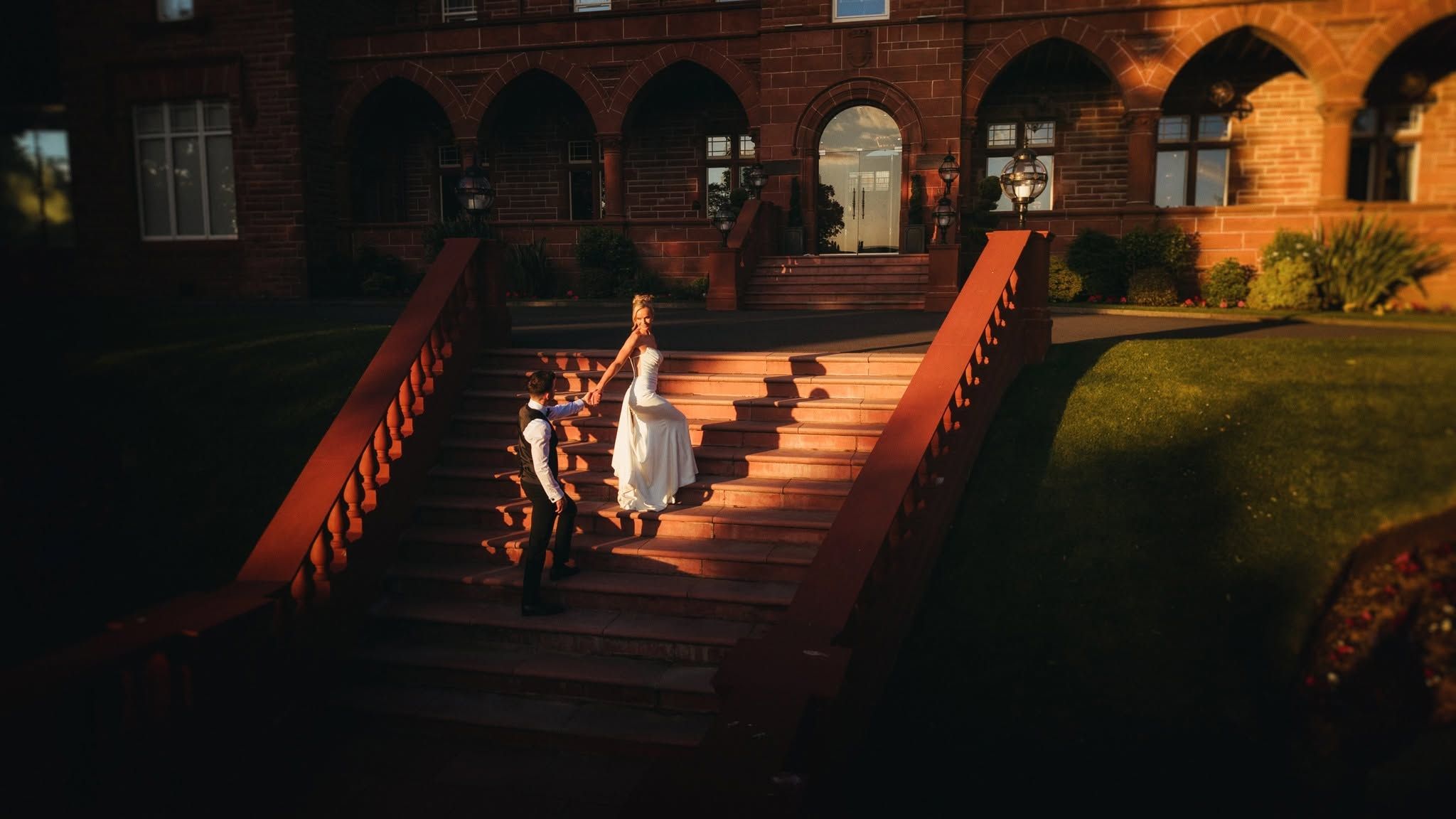 Bride in white dress descends ornate red brick castle stairs at golden hour sunset.