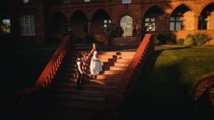 Bride in white dress descends ornate red brick castle stairs at golden hour sunset.