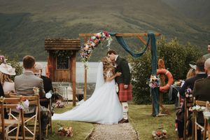 Bride and groom share first kiss at outdoor Scottish Highland wedding ceremony with floral arch and mountain backdrop.