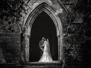Black and white wedding photo of bride and groom kissing in a Gothic stone archway surrounded by lush ivy.
