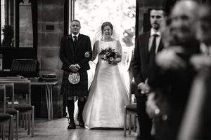 Black and white photo of a bride walking down the aisle in a white gown, holding a bouquet, escorted by a man in a kilt.