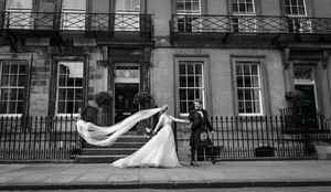 Black and white photo of a bride and groom outside a Georgian building, her veil flowing in the wind.