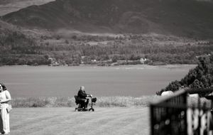 Person with stroller on green lawn overlooking scenic lake and mountain landscape in black and white photo.