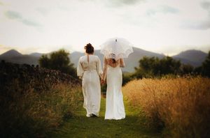Two brides in white gowns hold hands on a scenic path, one carrying a lace parasol, with mountains in the background.