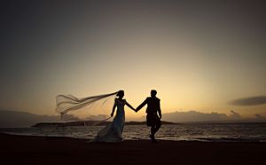 Silhouette of a bride and groom holding hands on a beach at sunset, with the veil flowing in the wind.