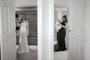 Black and white wedding photo of bride in lace gown while woman holds baby in adjacent room.