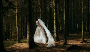 A bride in a flowing white wedding gown and veil twirls gracefully among tall trees in a dark forest.