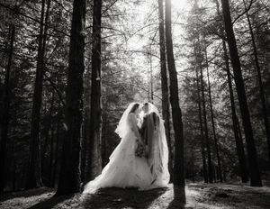 Black and white photo of two brides sharing a romantic moment in a sunlit forest wearing elegant wedding gowns and veils.