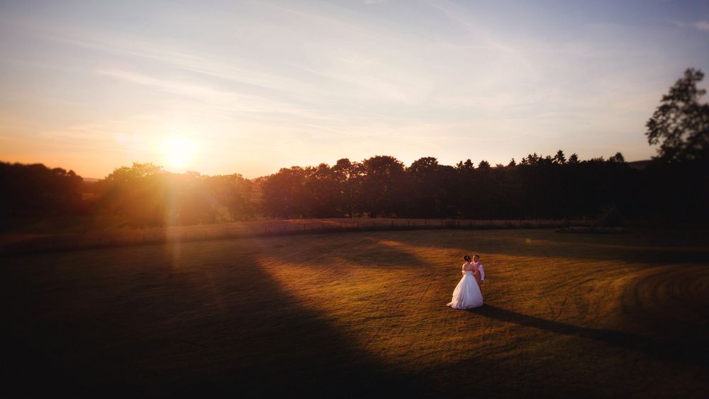 Bride and groom embracing at sunset on a lush green field with warm golden light and tree silhouettes.