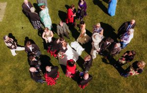 Aerial view of wedding guests gathered on green lawn, wearing formal attire in colorful outfits, celebrating outdoors.