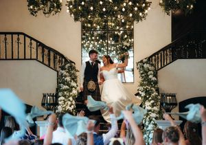 Bride and groom stand on grand staircase adorned with greenery and lights as guests wave ribbons below.
