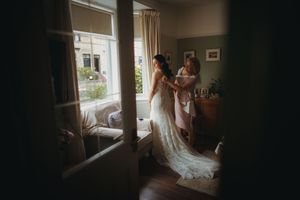 A bride in a lace wedding gown being helped by a bridesmaid near a window in a softly lit room.