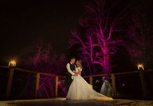 Bride and groom share romantic kiss on outdoor deck at night with pink lit trees glowing behind them.