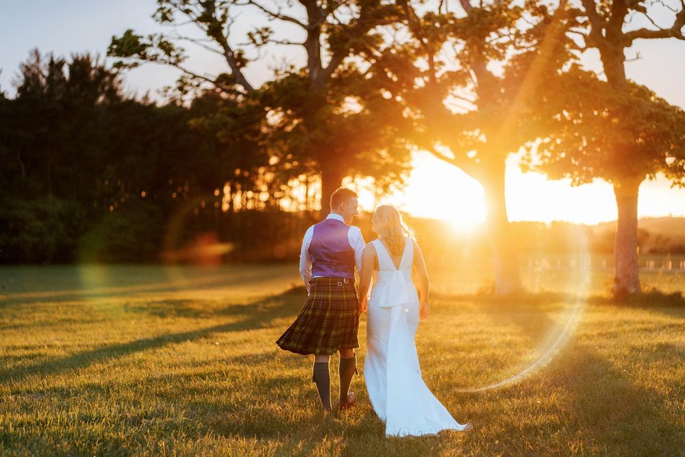 Bride and groom walk hand-in-hand through golden sunset light in a lush green field, man wearing traditional Scottish kilt.