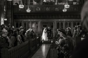 Black and white photo of a wedding ceremony in an ornate church, bride and groom at the altar with guests seated in pews.
