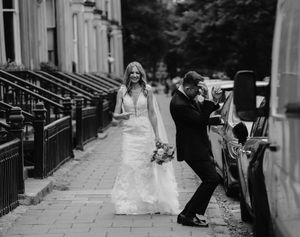 Black and white photo of a bride in a lace gown laughing as groom leans against a car on a city sidewalk.