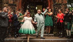 Joyful couple exits wedding venue in colorful attire, surrounded by guests throwing confetti outdoors.