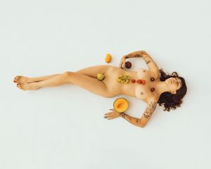 Artistic nude figure lying on white background with colorful fruits arranged on body in creative still life composition.