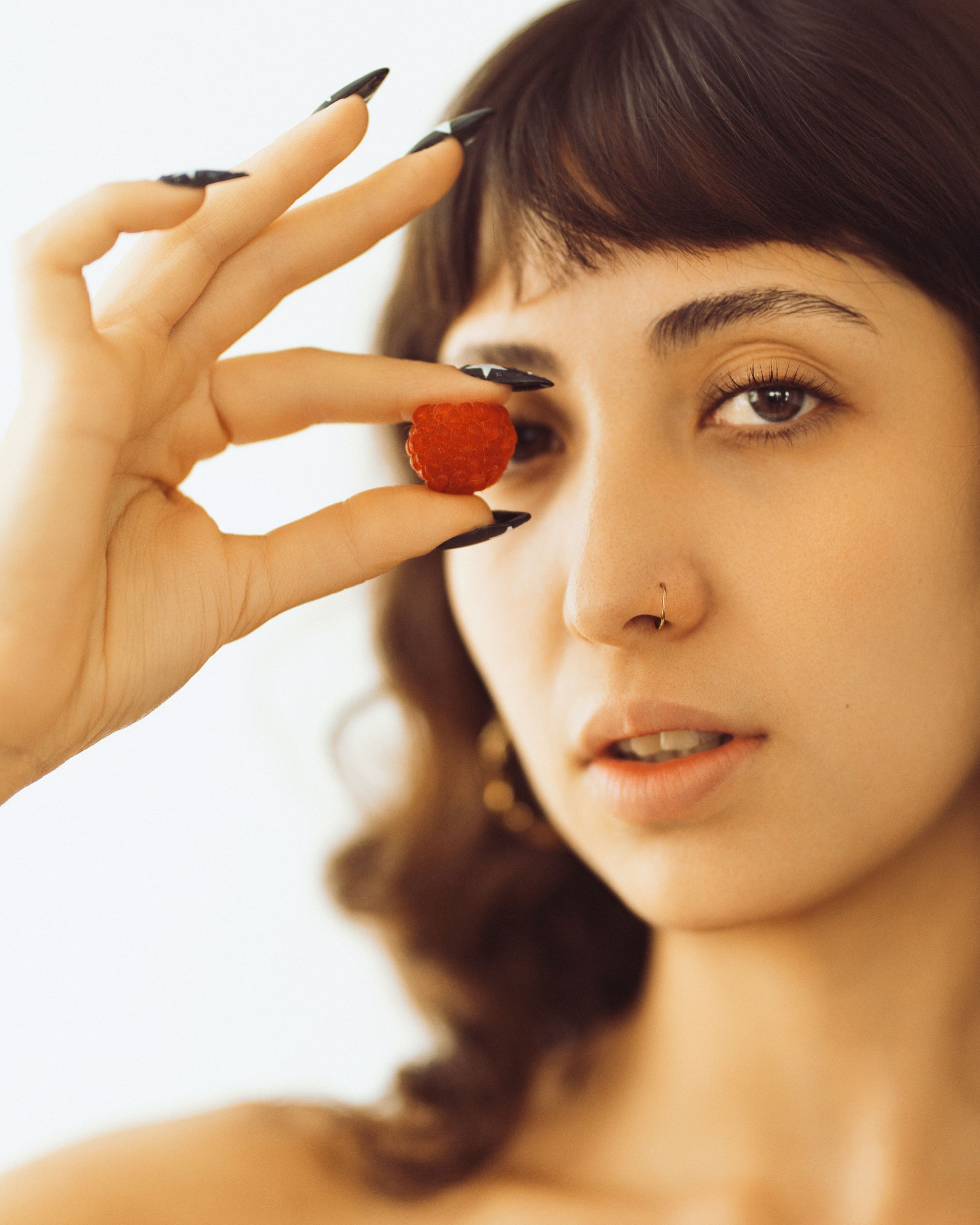 Woman with dark nails holds a red gemstone ring near her eye, wearing a nose ring, against white background.