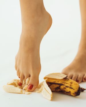 Bare feet with red toenails stepping on a ripe banana peel on a white background.