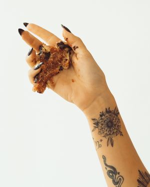 Tattooed hand with black nails holding a messy fried chicken piece, with floral and snake tattoos on the forearm.