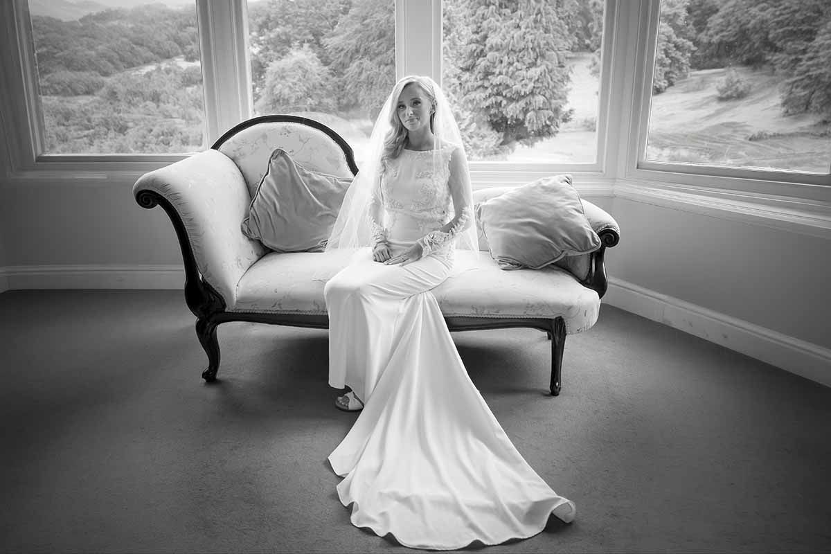 Bride in white gown and veil sits gracefully on vintage chaise lounge by large windows in black and white photo.