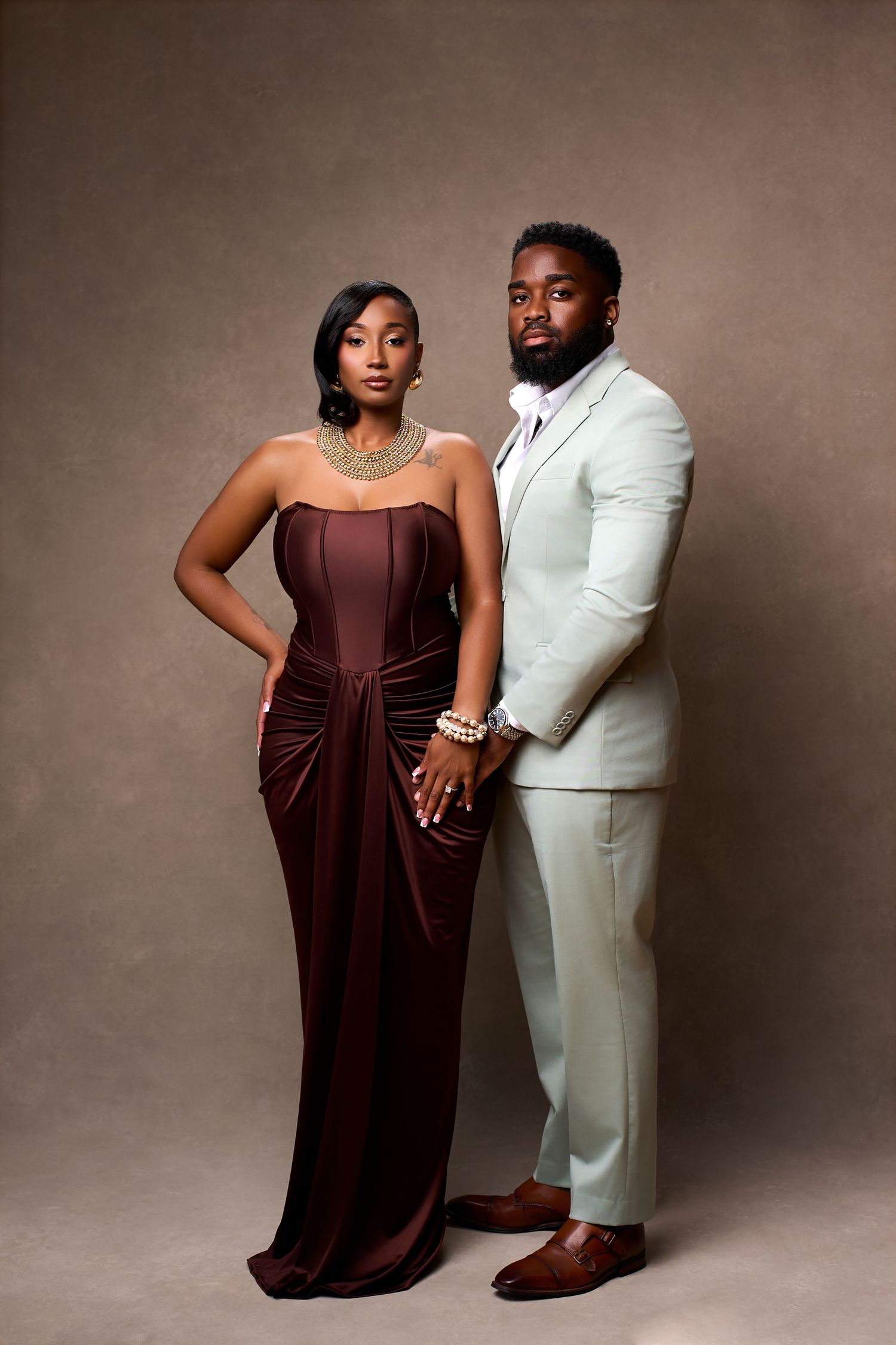 Elegant couple posing for their engagement shoot in studio, woman in burgundy gown with gold jewelry, man in cream suit with brown shoes.