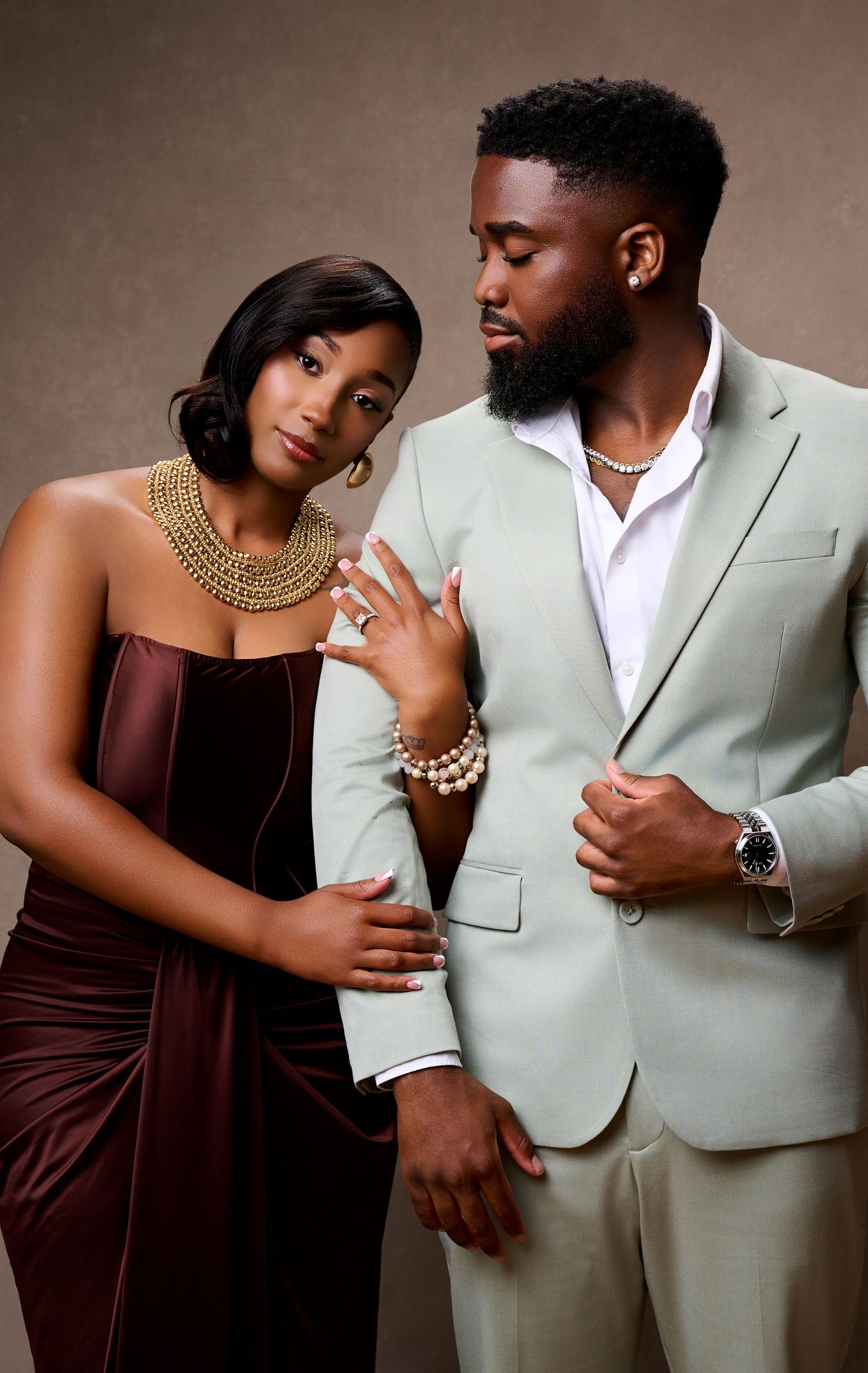 Elegant couple posing for their engagement in studio, woman in burgundy gown with gold jewelry, man in mint green suit with pearl necklace.