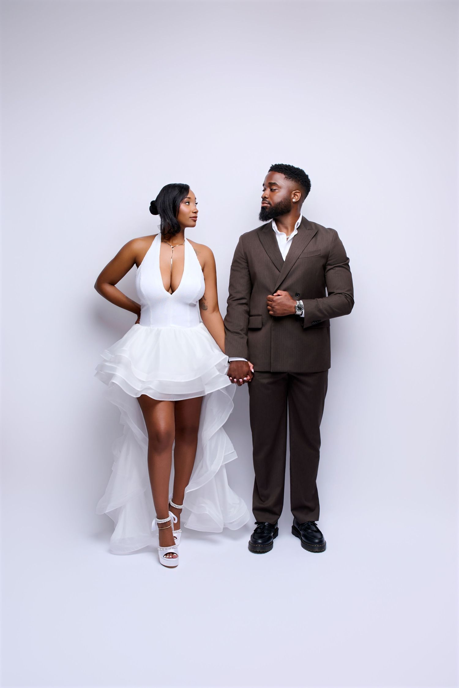 Black bride and groom holding hands in studio, she wears white halter mini dress, he wears a brown suit.