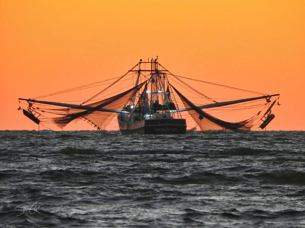 Commercial shrimp boat with outriggers extended silhouetted against a glowing orange sunset, photo by Steve Cole.