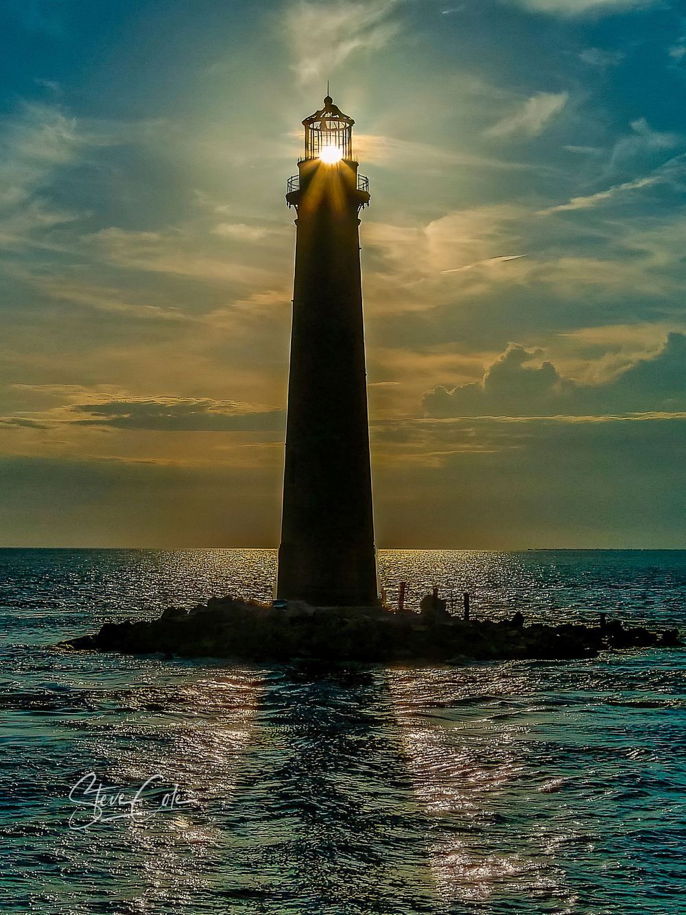 Silhouette of a historic lighthouse backset against the setting sun over ocean waters, photo by Steve Cole Photography.