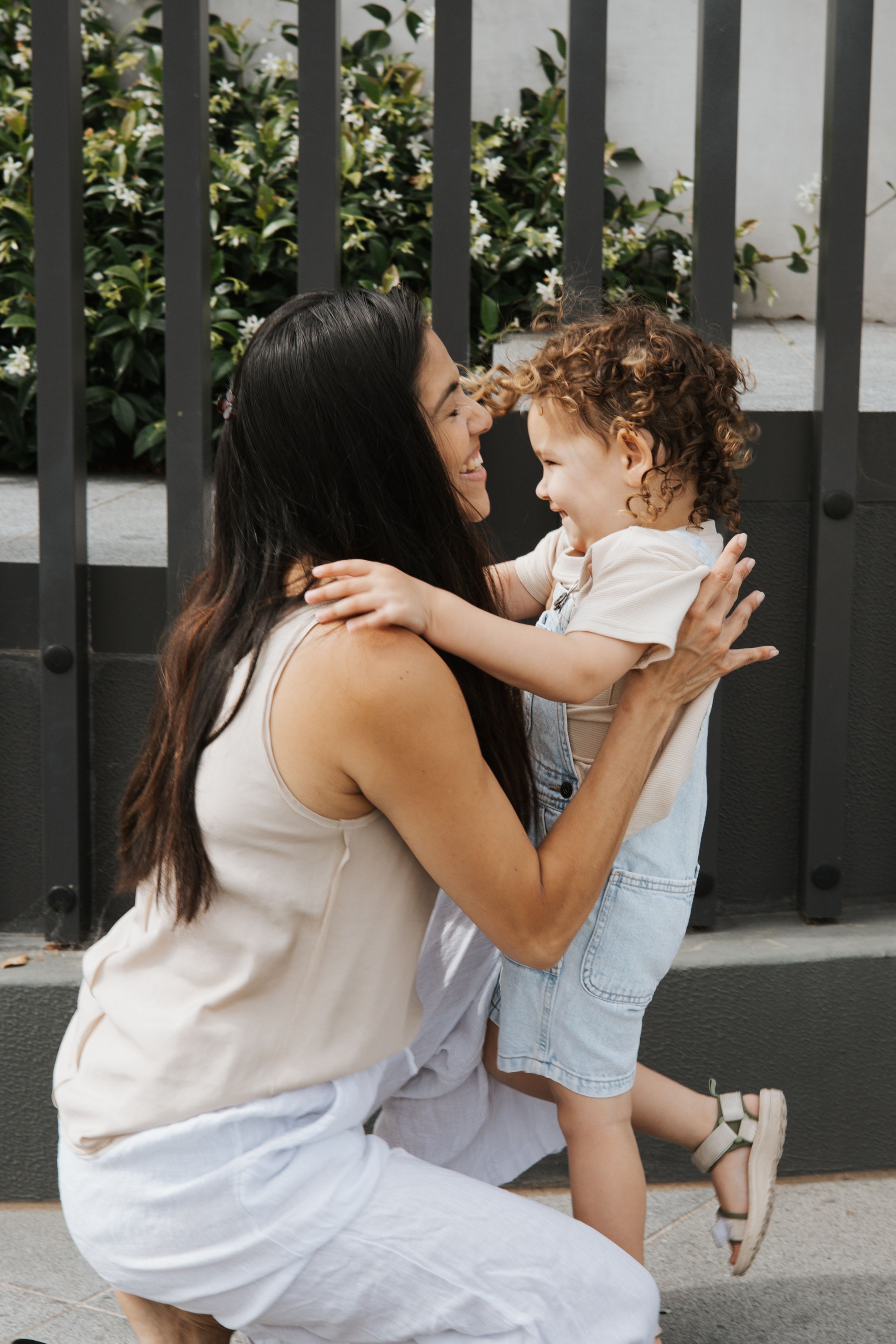 Dark-haired mother crouching to embrace curly-haired toddler outdoors near garden fence.