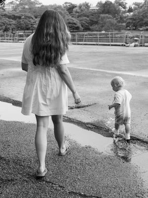 Black and white photo of a girl in white dress watching a toddler splash in a puddle on a paved path.